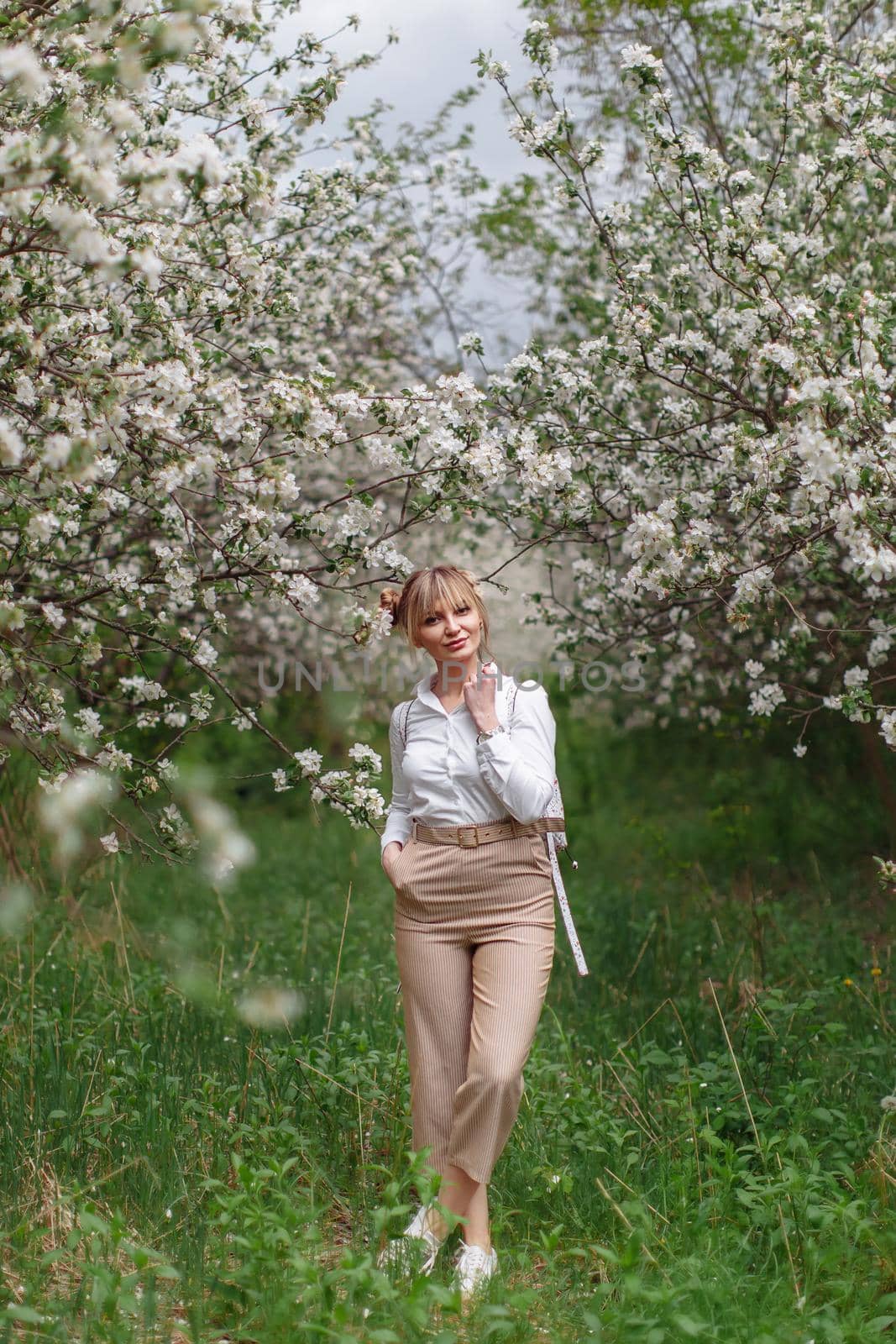 Beautiful young blonde woman in white shirt with backpack posing under apple tree in blossom in Spring garden by OnPhotoUa