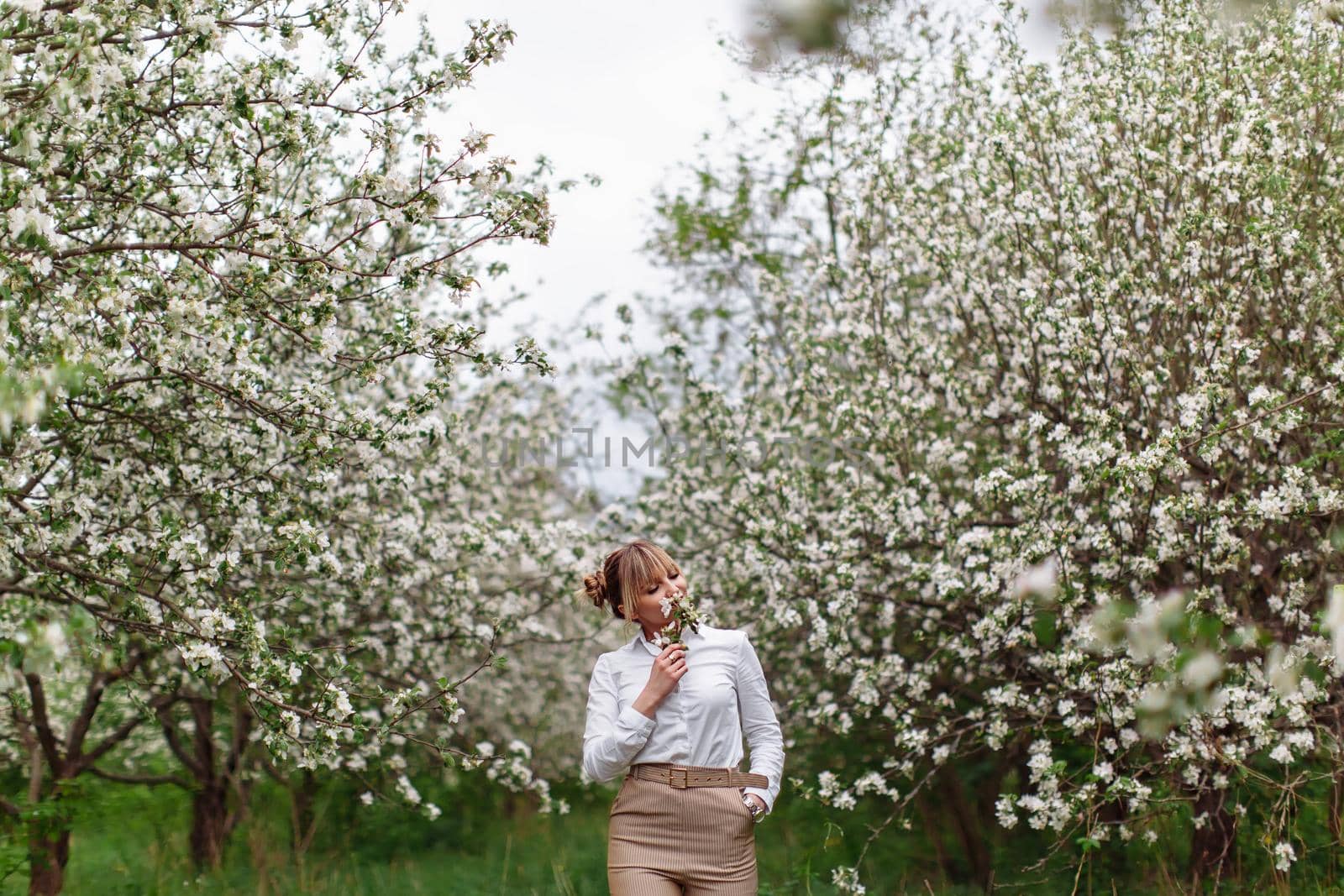 Beautiful young blonde woman in white shirt posing under apple tree in blossom and green grass in Spring garden