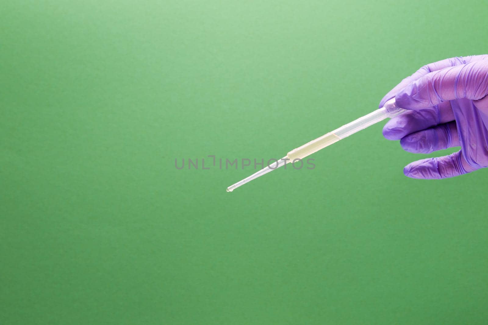 a female hand in a blue disposable glove holds a large plastic dropper, drips green liquid into a flask, green background, copy space by natashko