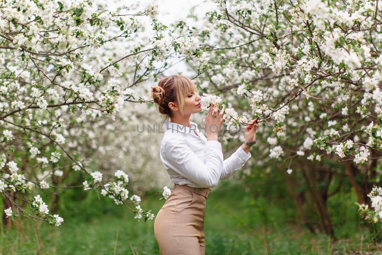 Beautiful young blonde woman in white shirt posing under apple tree in blossom in Spring garden by OnPhotoUa