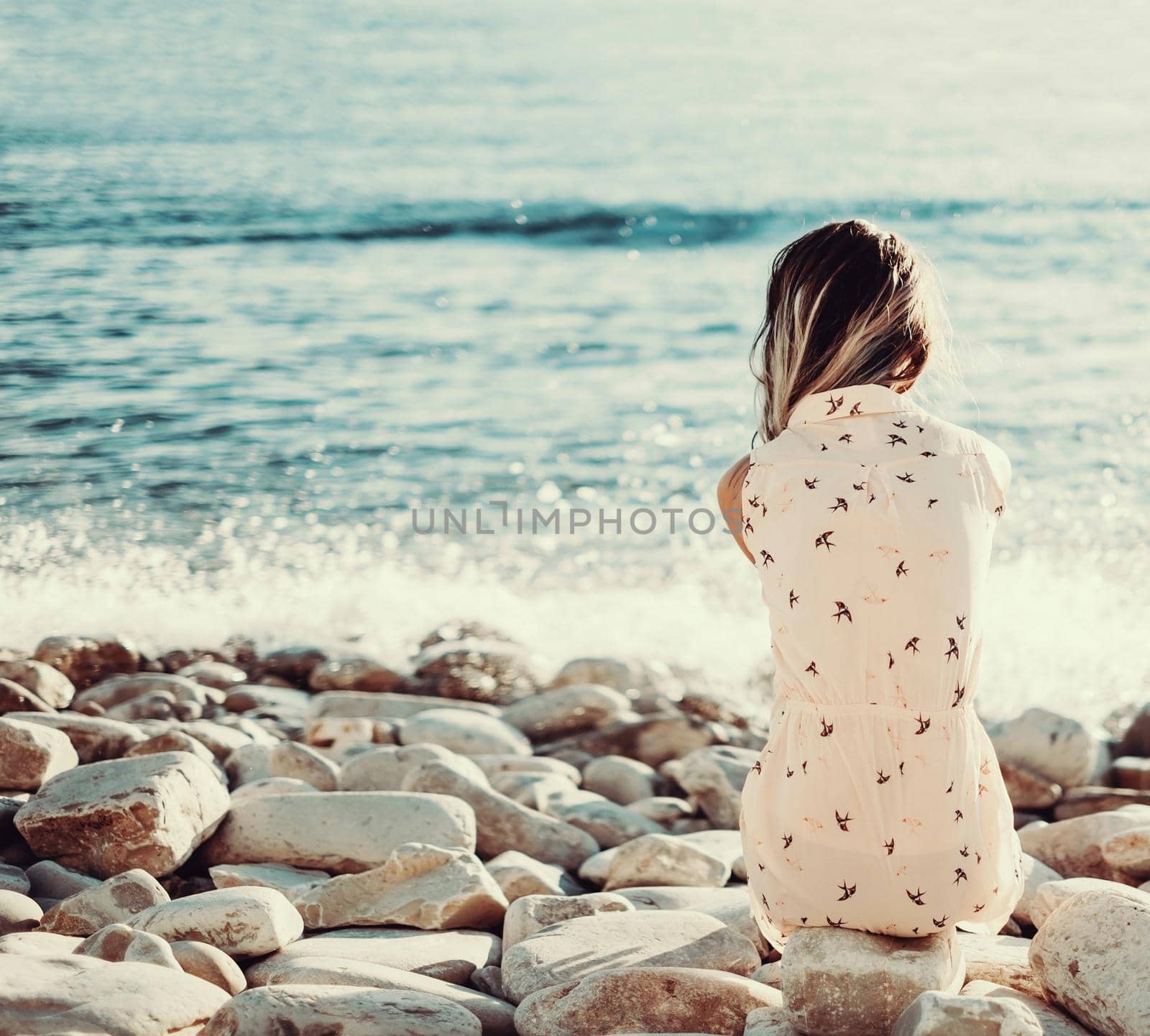 Young woman resting on pebble coast and enjoying view of sea, rear view. Toned image. Space for text in left part of image.