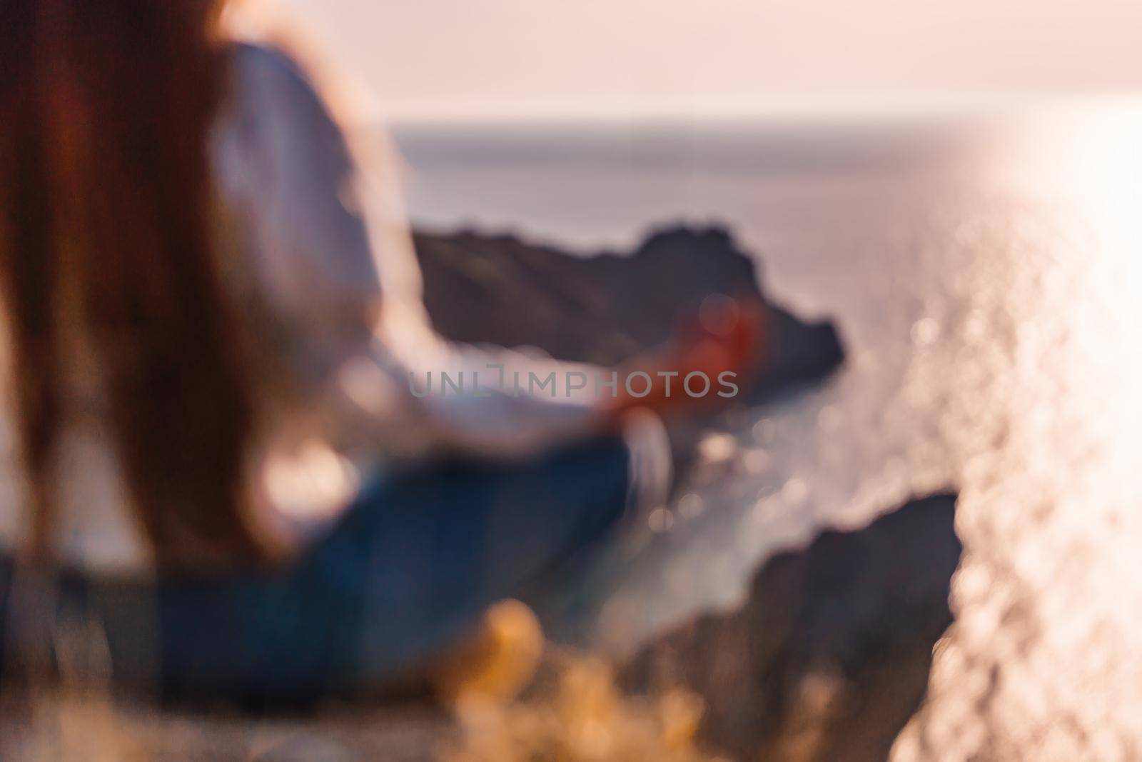 Defocused silhoute of Young woman with long hair in sportswear practicing outdoors on yoga mat by the sea on a sunset. Women's yoga fitness routine. Healthy lifestyle, harmony and meditation by panophotograph