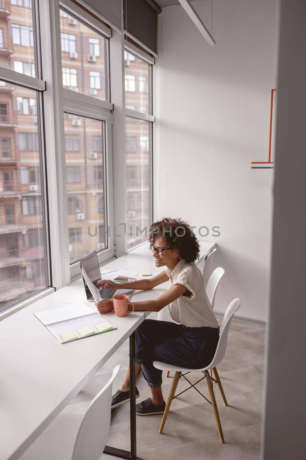 Smiling Afro American businesswoman using laptop and holding report while sitting near the window