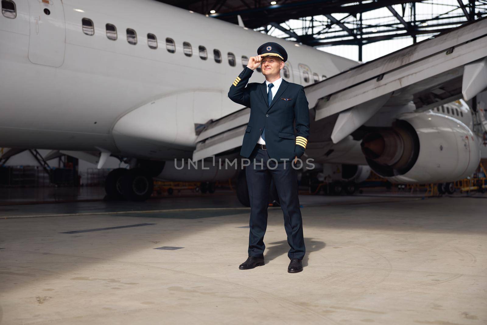 Full length shot of confident pilot in uniform looking away, adjusting his hat, standing in front of big passenger airplane in airport hangar. Aircraft, occupation, transportation concept