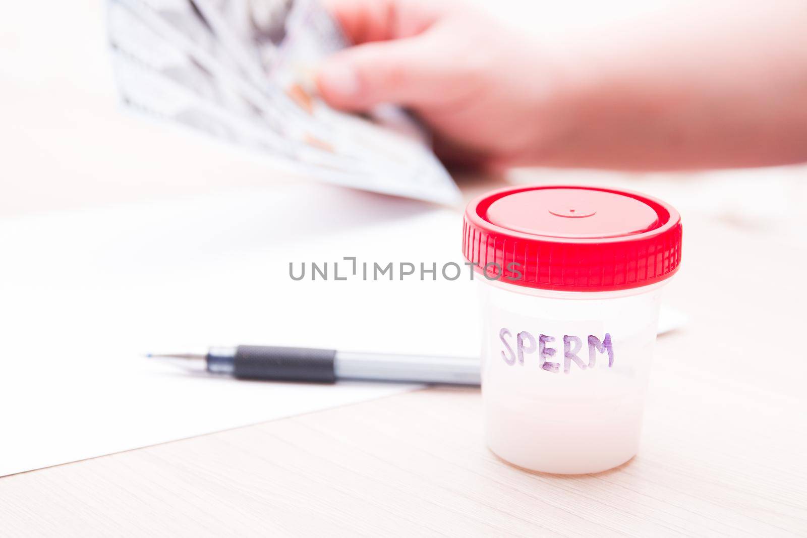 test jar with a red lid and the inscription sperm, against the background a man holds several notes of 100 dallors, a copy space, sperm in a jar, a sperm donor concept, sell sperm, a pen and paper on the table