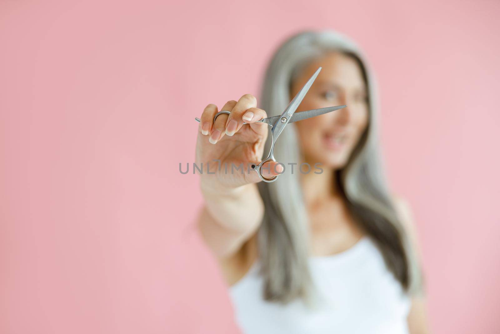 Happy middle aged Asian woman poses on pink background, focus on hand with scissors by Yaroslav_astakhov