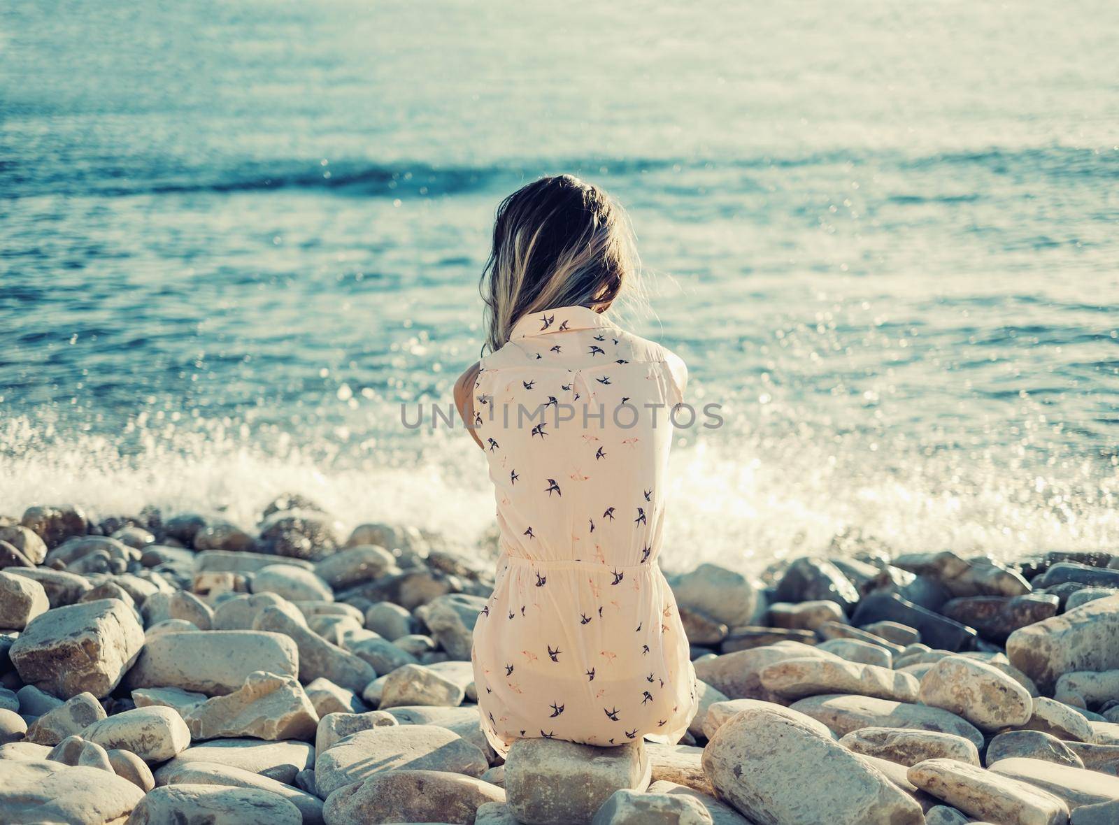 Young woman resting on pebble beach and enjoying view of sea, rear view. Toned image.