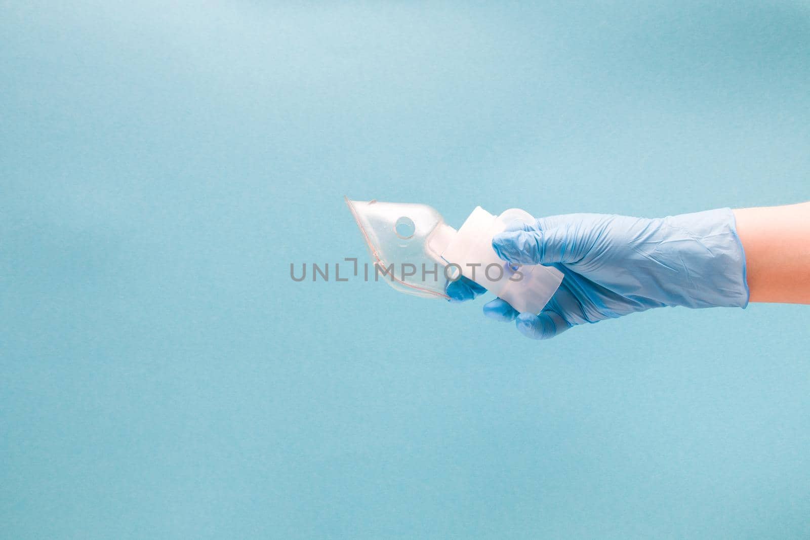 a female hand in a blue disposable medical glove holds a nebulizer medication reservoir with a baby mask, inhalation device, respiratory tract disease concept, pulmonology and respiratory infections treatment, blue background, copy space
