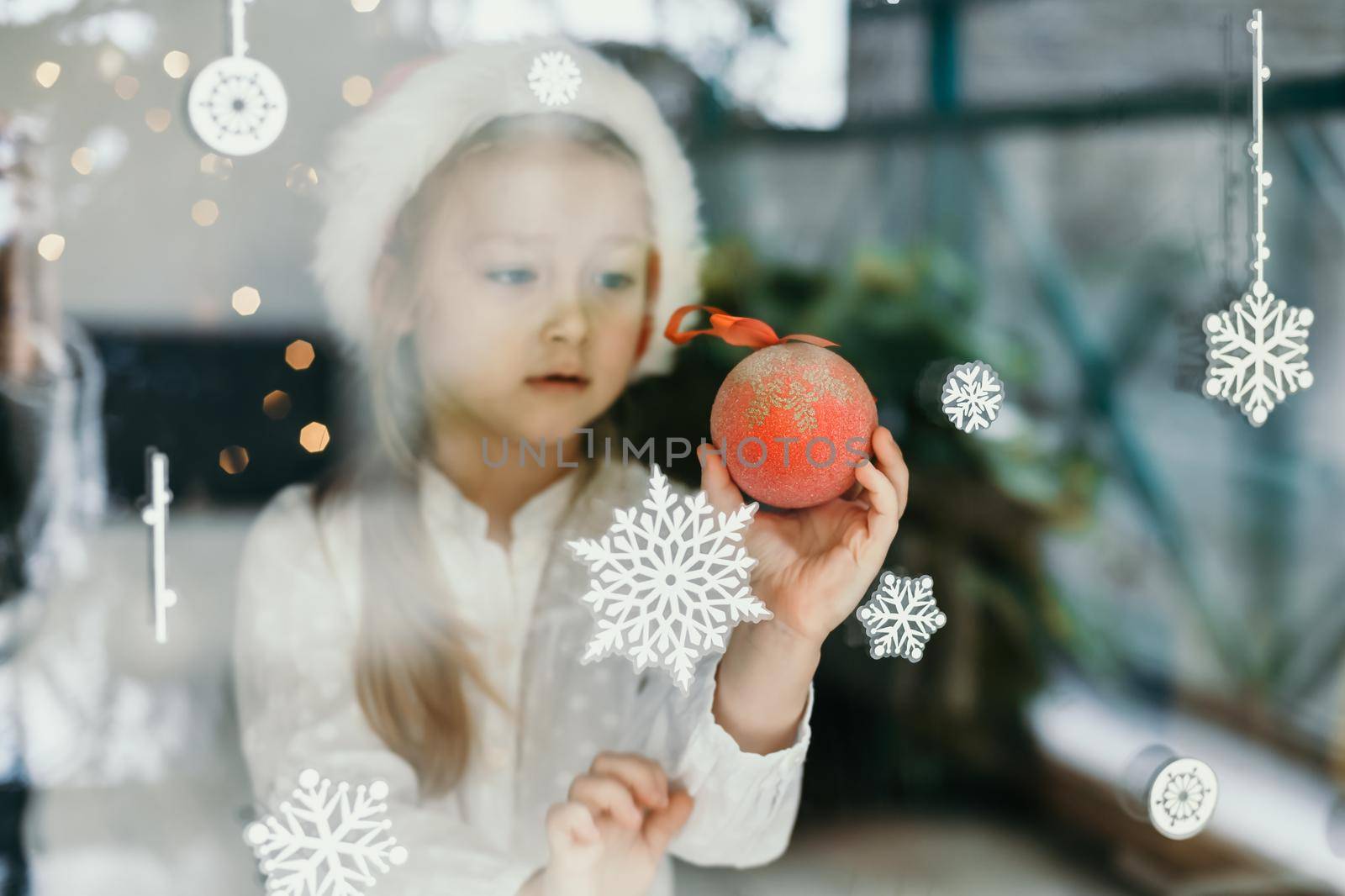 A girl wearing a santa claus hat looks at snowflakes on a window