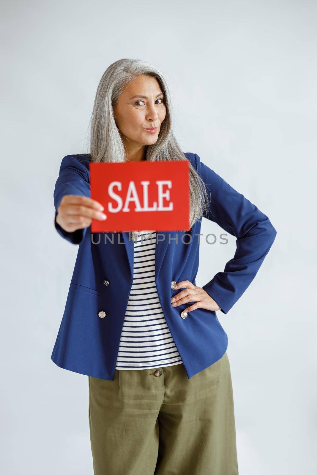 Cute mature Asian lady with loose grey hair wearing elegant jacket holds red card with word Sale posing on light background in studio