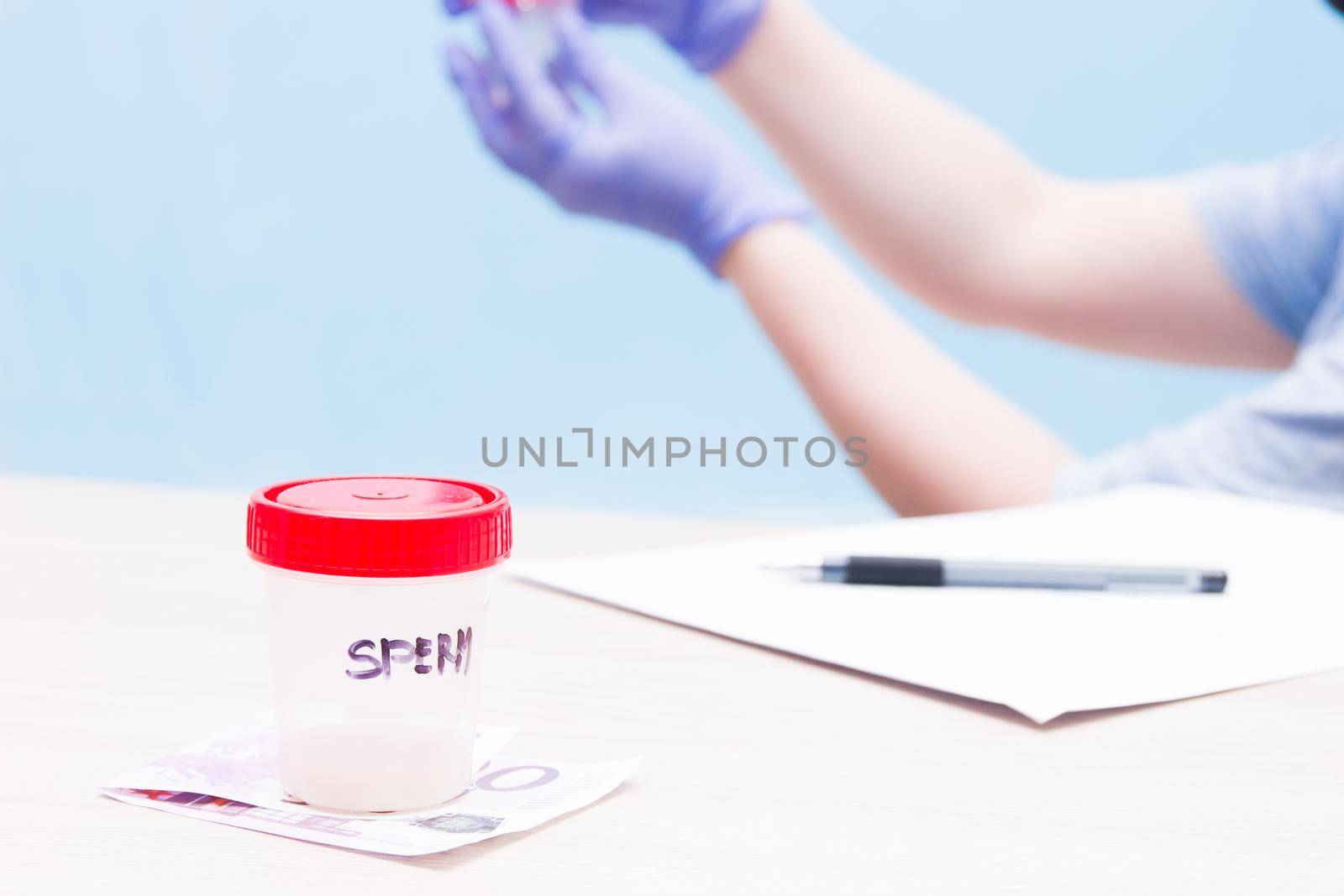 a jar with a red lid with sperm, stand on a banknote of 500 euros, a sperm donor concept, female hands with a jar for analysis on a blue pan in blue gloves, blue background, copy space, paper and pen on the table