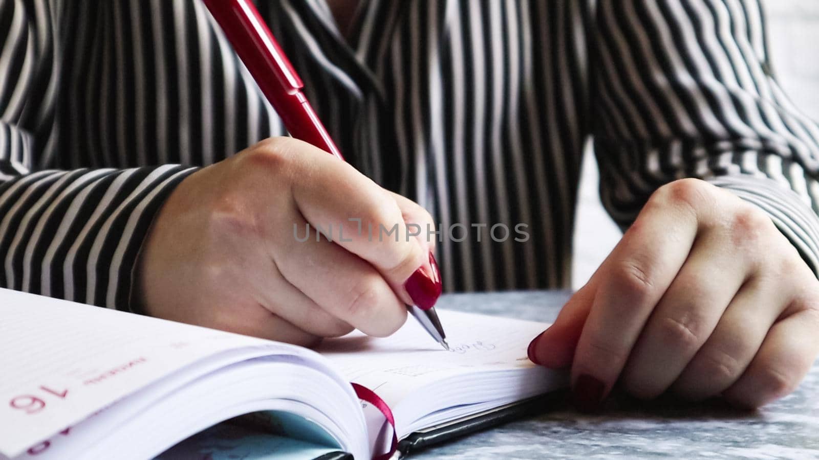 Side view of the hands of a woman with red nails, holding a red pen, writes something on an empty sheet of diary paper lying on a gray table. Girl makes notes in a black notebook.