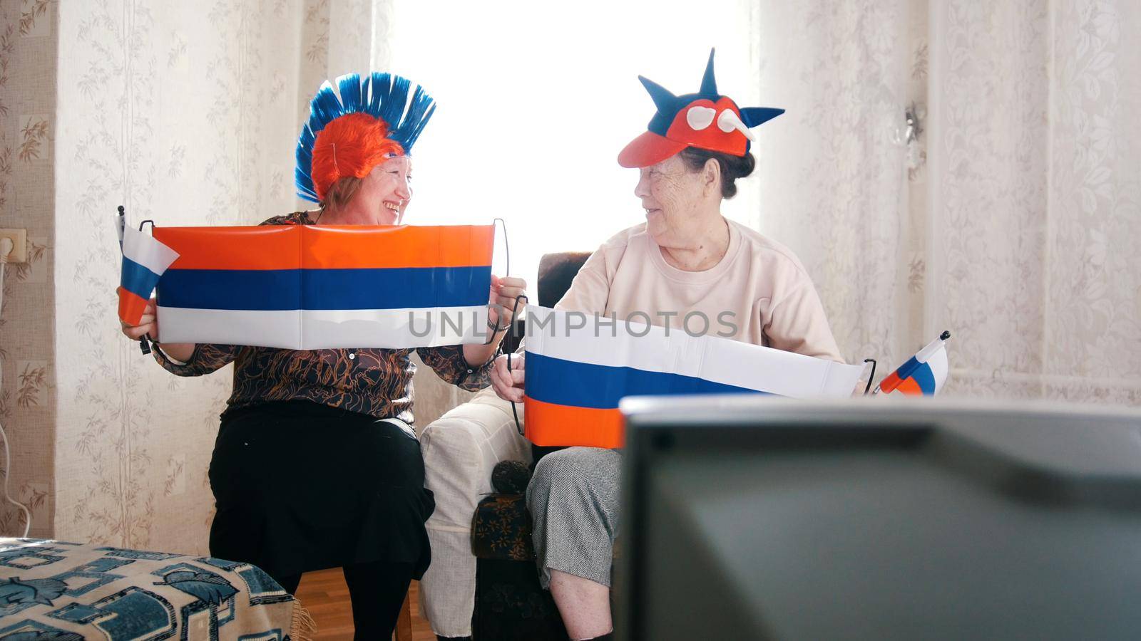 Two elderly women watching in russian accessories TV. Russian flags. Portrait