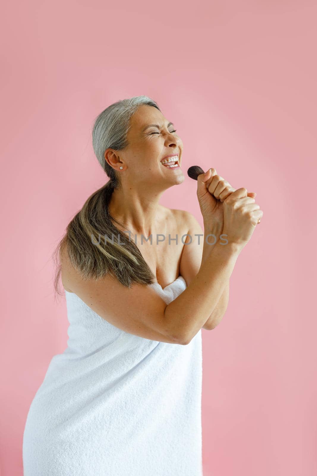 Happy silver haired Asian lady sings song using brush as microphone standing on pink background in studio. Mature beauty lifestyle