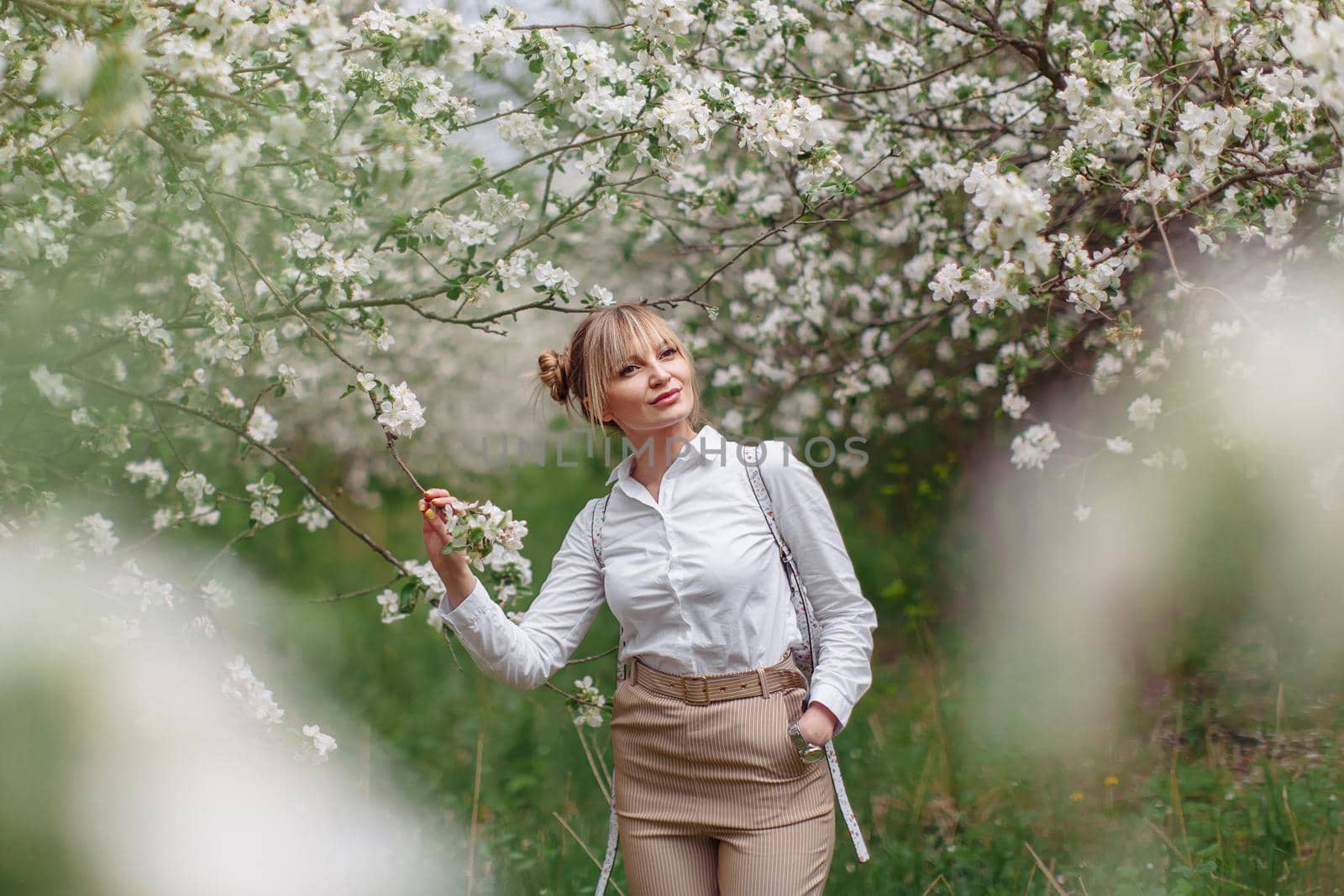 Beautiful young blonde woman in white shirt with backpack posing under apple tree in blossom in Spring garden by OnPhotoUa