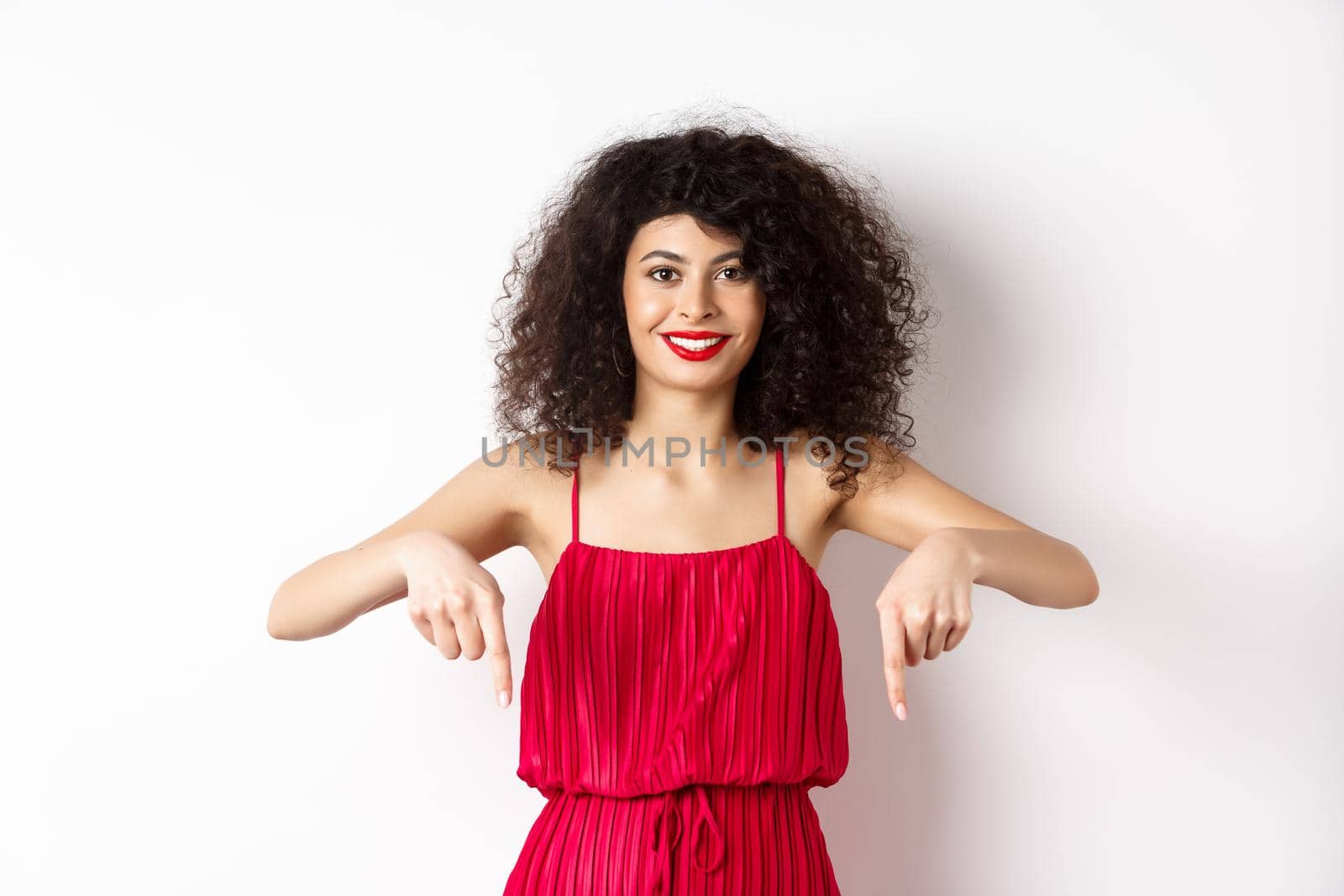 Attractive female model in red dress and makeup, pointing fingers down and smiling, showing advertisement, standing over white background by Benzoix