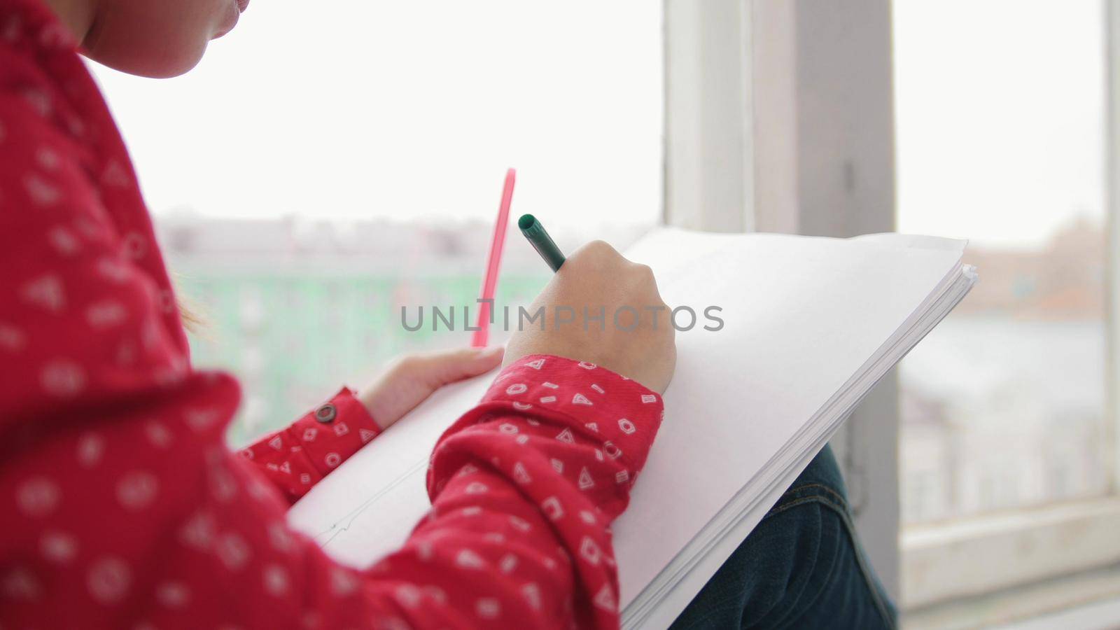 A little girl sitting on the window sill and drawing, smiling on the backround of the city. Close up photo. Portrait