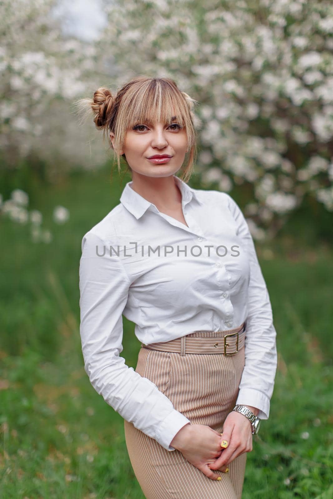 Beautiful young blonde woman in white shirt posing under apple tree in blossom and green grass in Spring garden