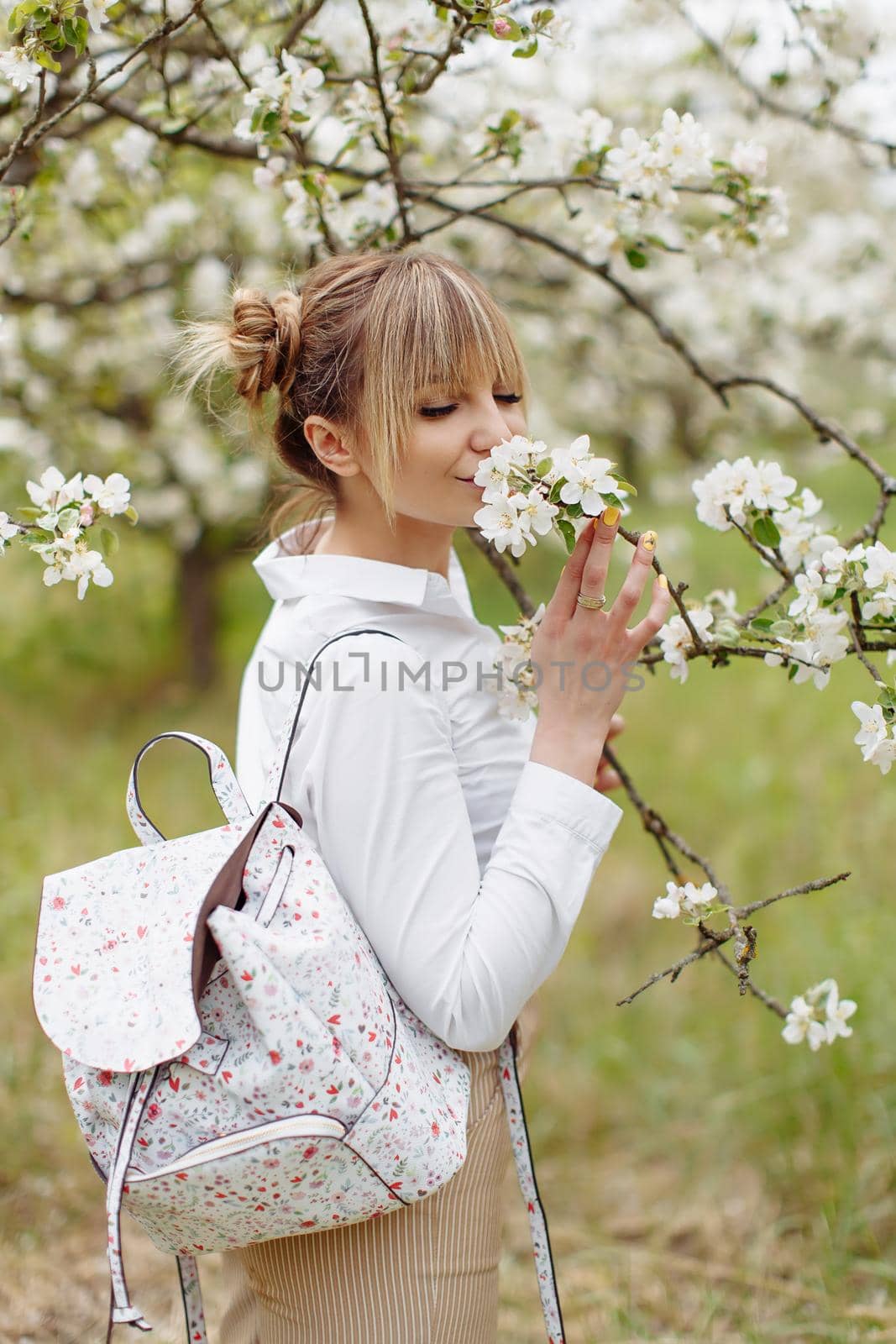 Close-up portrait of beautiful young blonde woman in white shirt with backpack posing under apple tree in blossom and green grass in Spring garden