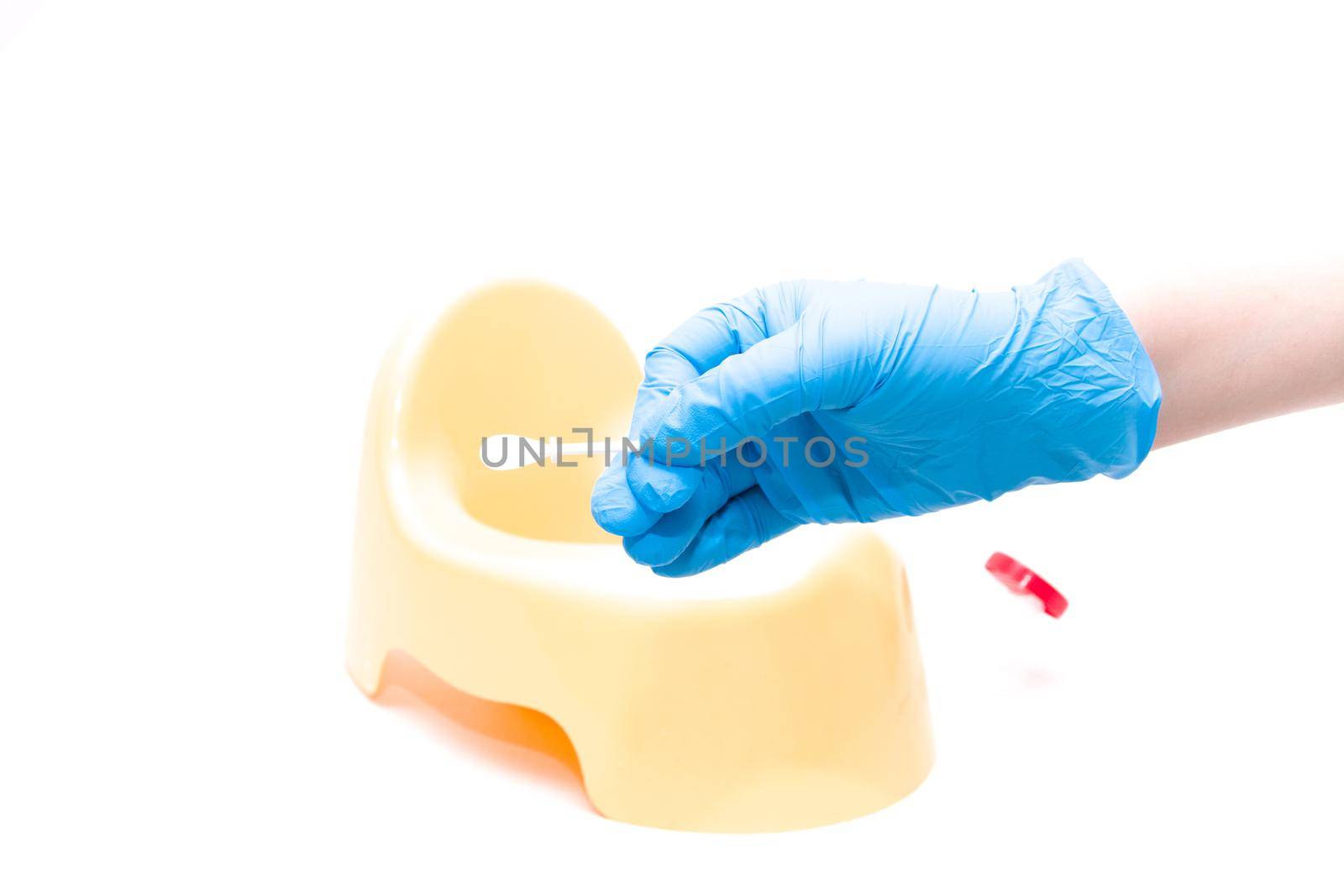 a hand in a disposable blue medical glove holds a test jar with a white spoon for collecting feces, in the background is a yellow children's pot, white background, copy space by natashko