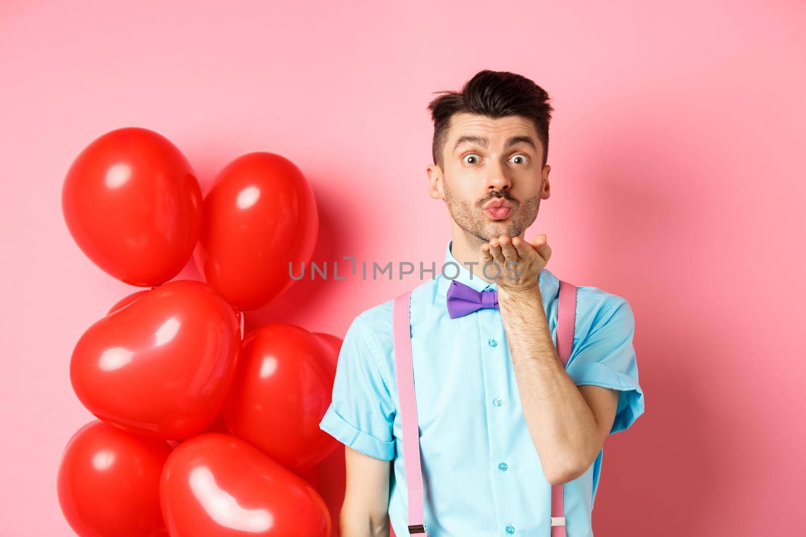 Valentines day concept. Attractive and funny man in bow-tie, blowing air kiss to lover, standing near pink background and red hearts balloons by Benzoix