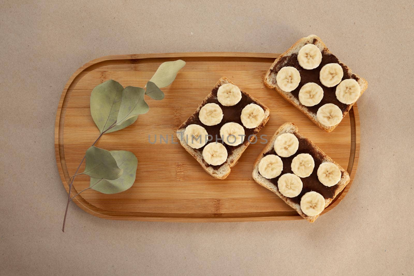 Three banana white bread toasts smeared with chocolate butter that lie on a cutting board with a sprig of leaves on craft paper background. top view with area for text