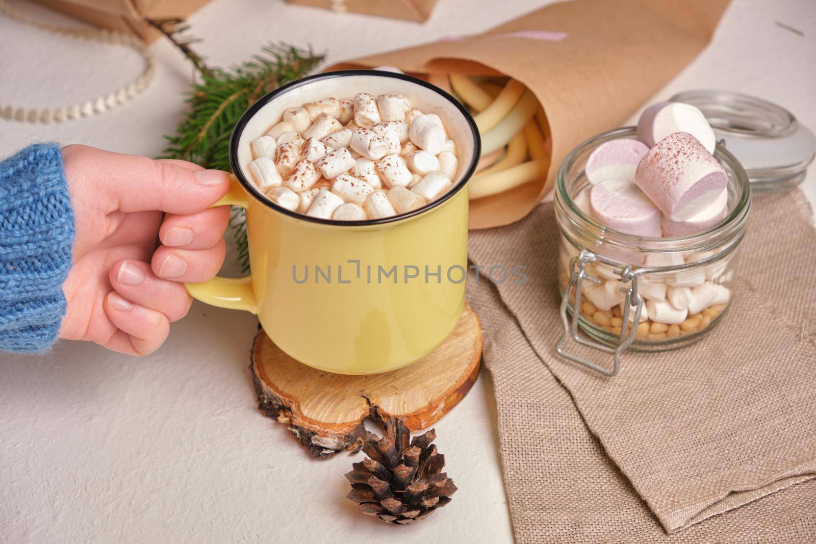 a hand in a sweater holds a yellow cup with cocao decorated with marshmallows, a stand made of saw cut wood and marshmallows on the background