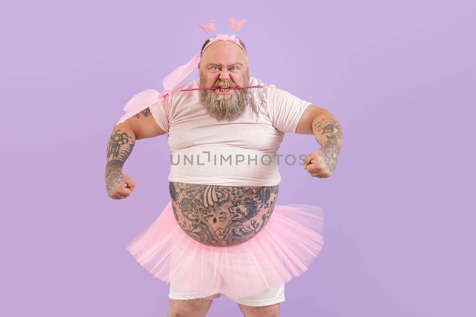 Funny brutal obese man with different tattoos in fairy costume with wings holds magic stick in teeth on purple background in studio