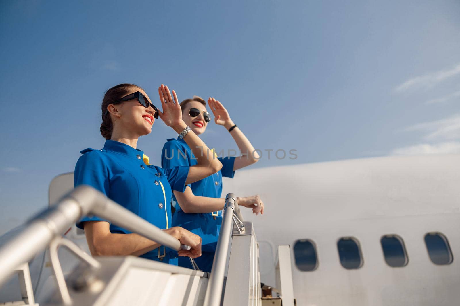 Portrait of two elegant air stewardesses in blue uniform and sunglasses covering eyes with hand and looking far away, standing together on airstair. Aircrew, occupation concept