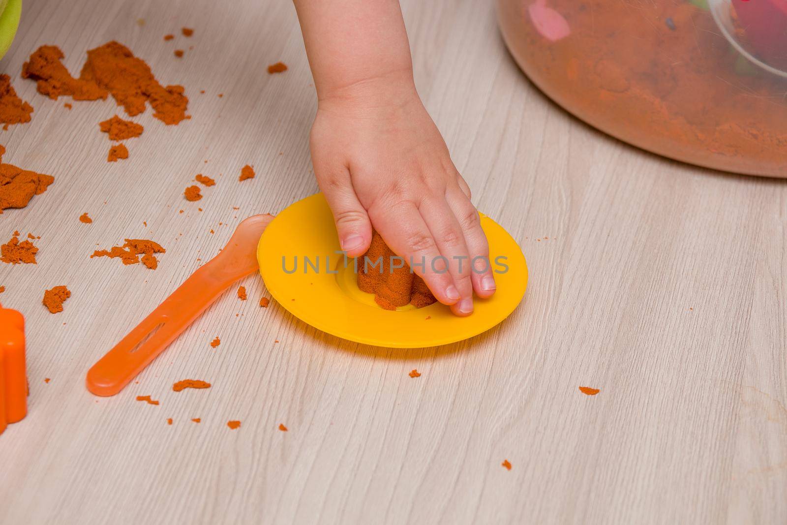 child's hand sculpts a cupcake on a yellow plastic orange sand toy plate on a wooden beige table