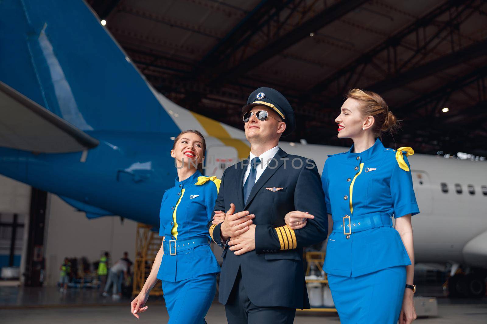 Happy pilot in uniform and aviator sunglasses walking together with two air stewardesses in blue uniform in front of big passenger airplane in airport hangar. Aircraft, occupation concept