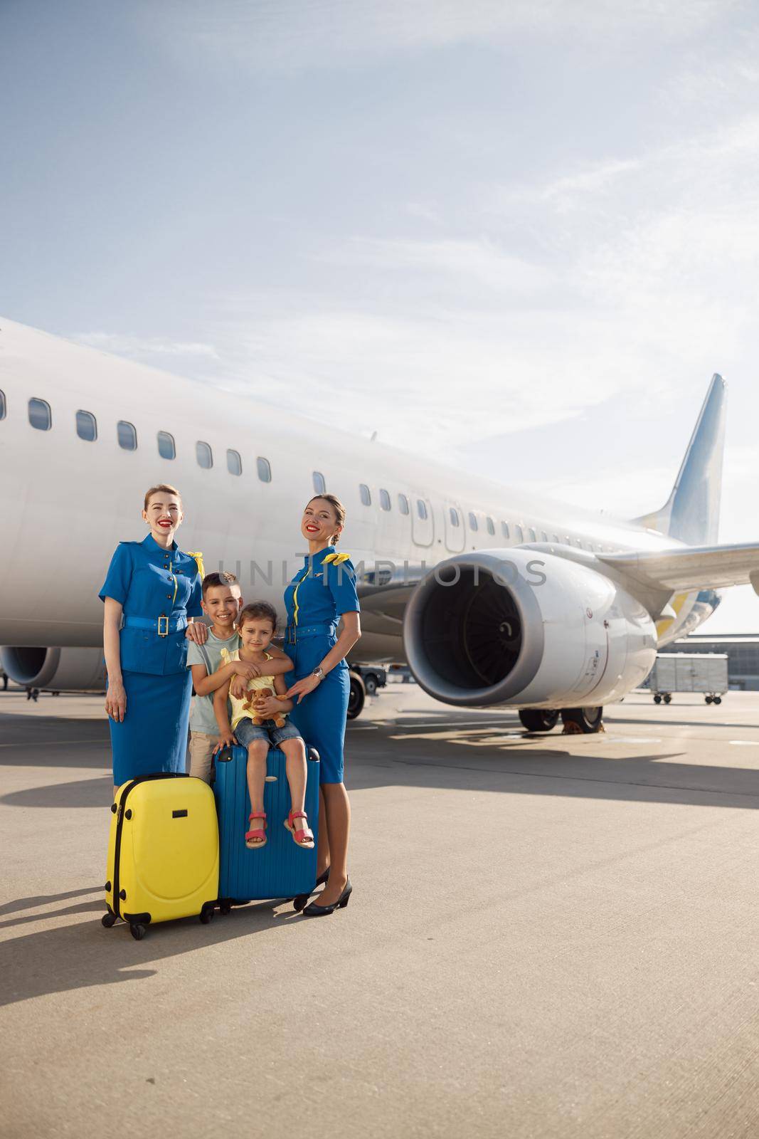 Full length shot of beautiful stewardesses standing near two kids sitting on their luggage in front of big airplane and smiling at camera by Yaroslav_astakhov