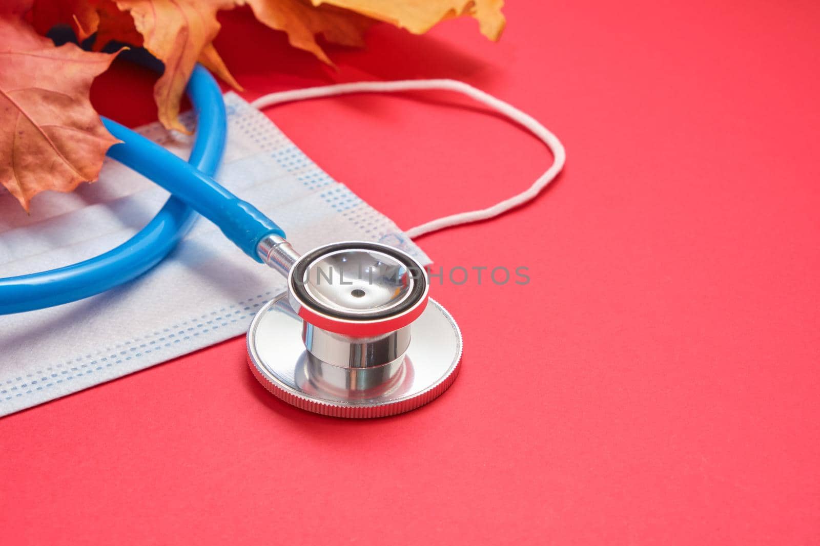 disposable protective medical mask and stethoscope and autumn maple leaves on a red background copy space, seasonal colds concept, coronavirus pandemic 2020 by natashko