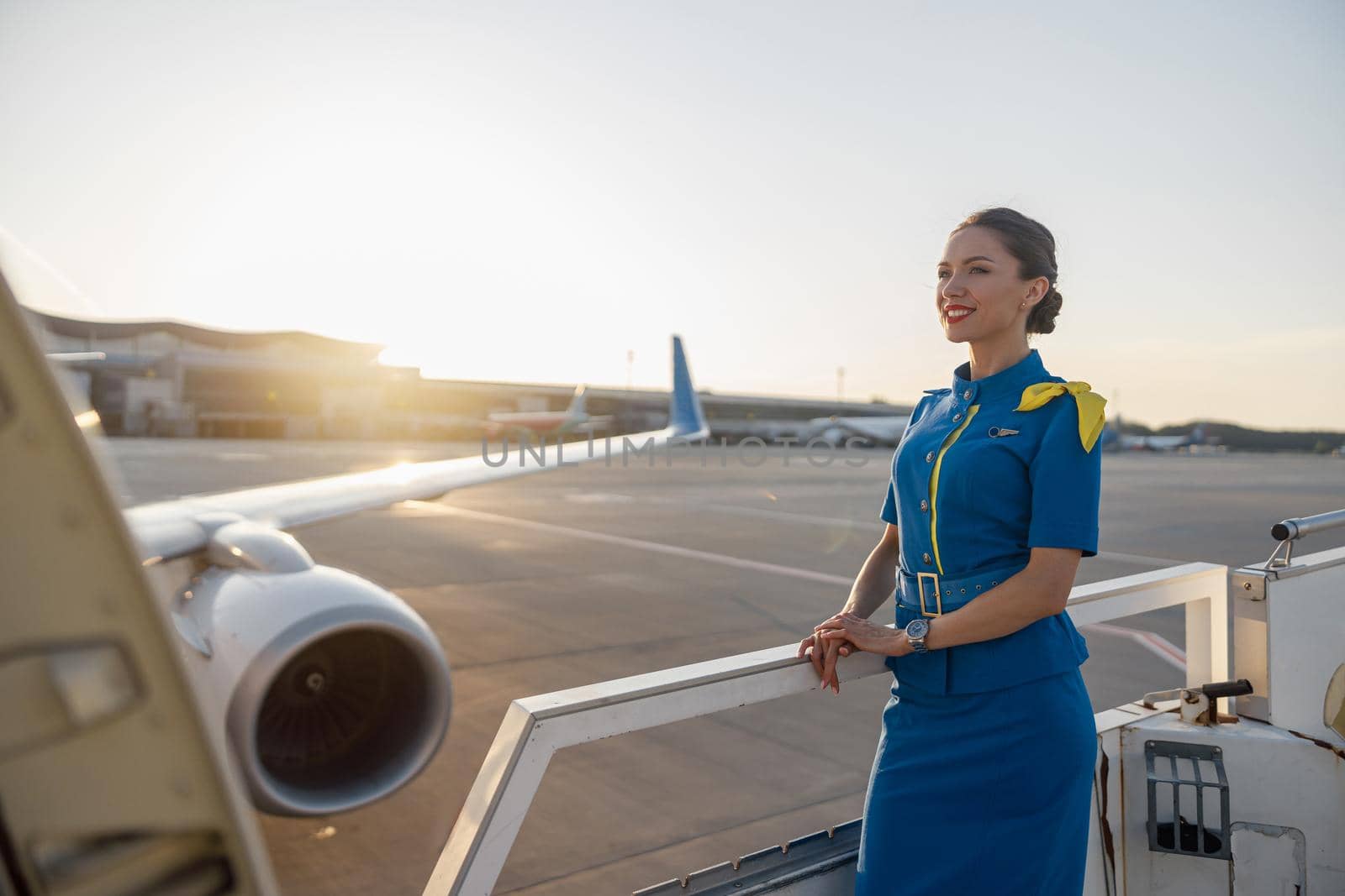 Pretty air stewardess in blue uniform smiling away, standing outdoors at the sunset. Commercial airplane near the terminal in an airport in the background. Aircrew, occupation concept