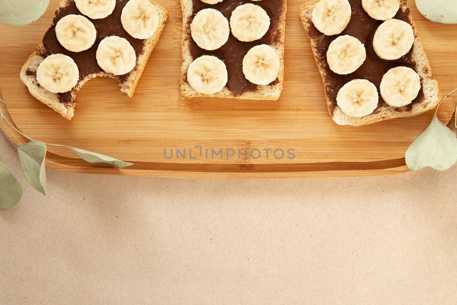 Three banana white bread toasts smeared with chocolate butter that lie on a cutting board with a sprig of leaves on craft paper background. top view with area for text