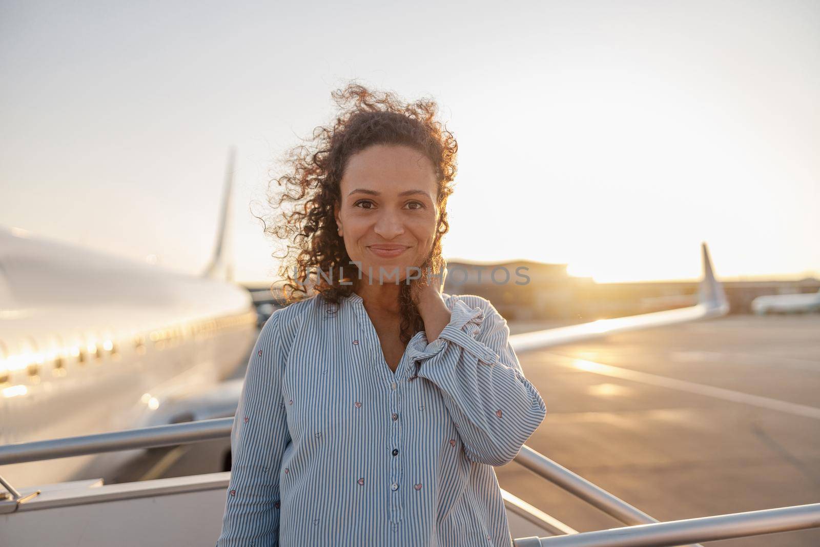 Portrait of relaxed woman smiling at camera while standing outdoors ready for boarding the plane at sunset by Yaroslav_astakhov
