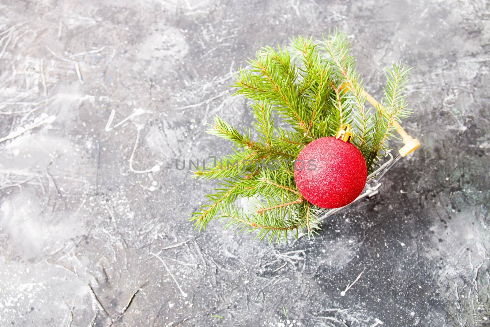 spruce green branch and red shiny Christmas-tree toy ball in a miniature shopping trolley on a black background, copy space by natashko