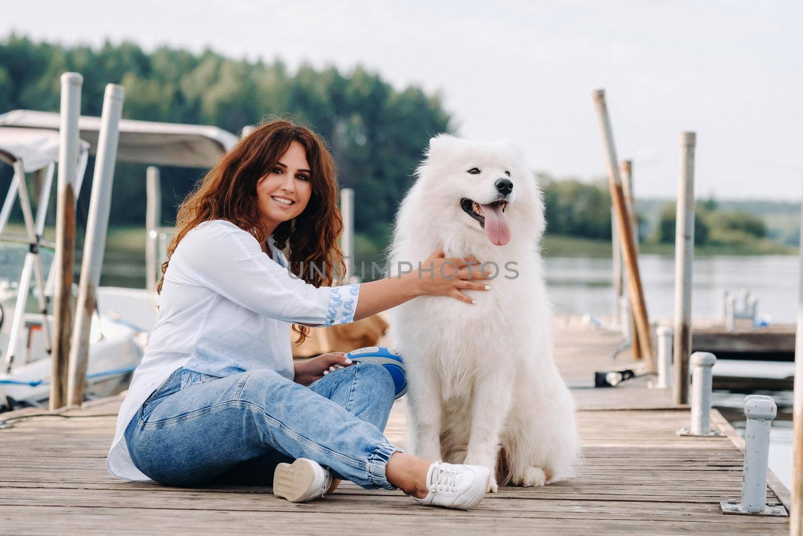 a happy woman with a big white dog lies on a pier near the sea at sunset by Lobachad