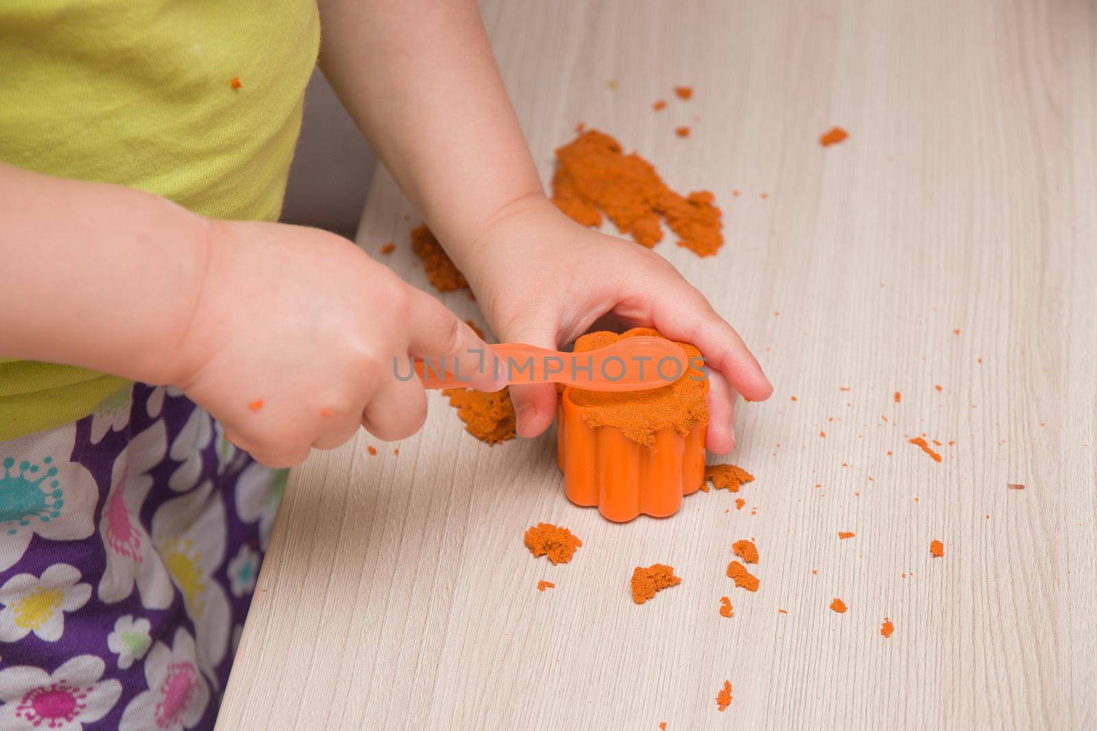 children's hand presses orange sand in a mold for toy muffins and cakes on a beige wooden table by natashko
