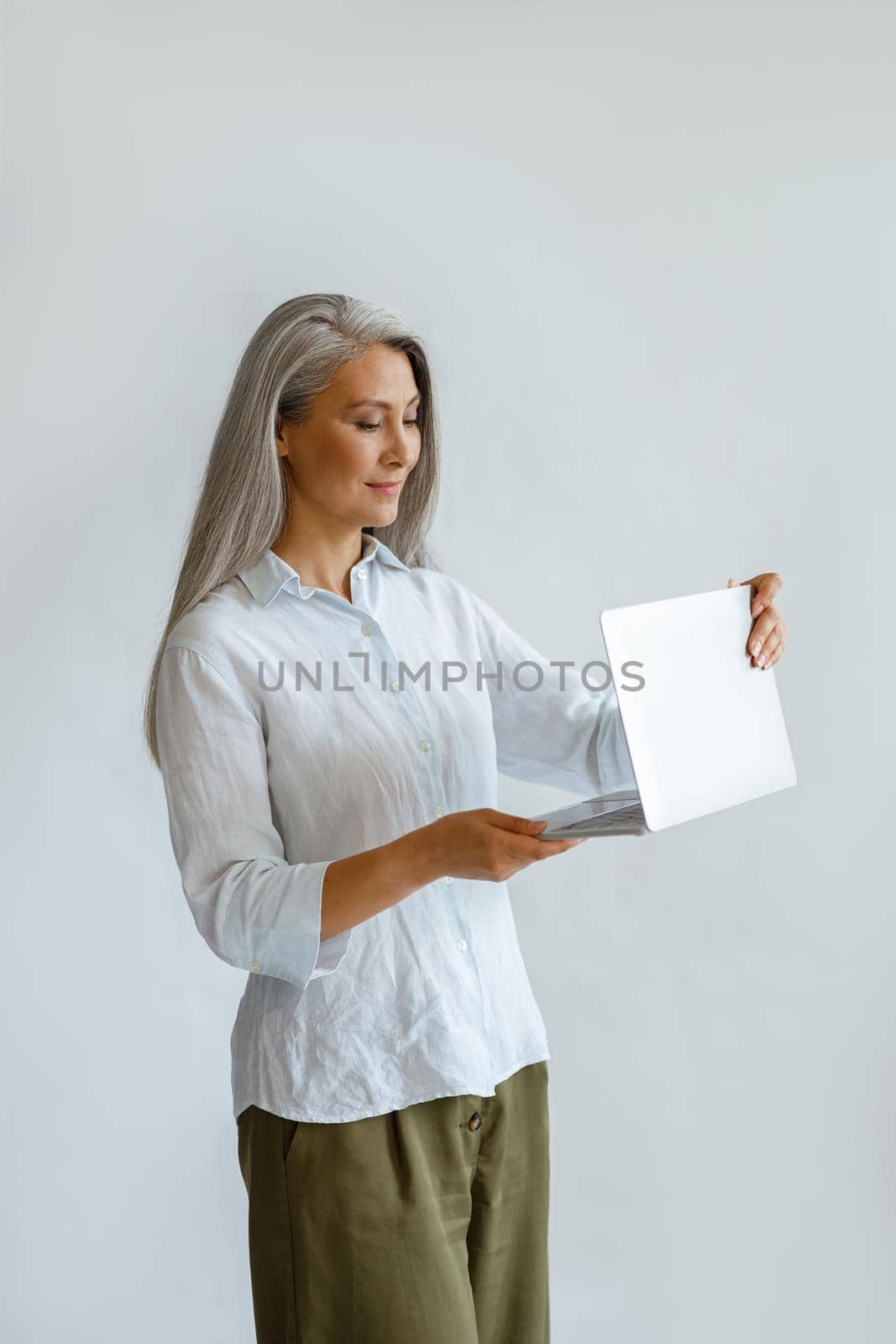 Happy mature Asian lady with hoary hair wearing casual clothes opens contemporary laptop standing on light grey background in studio