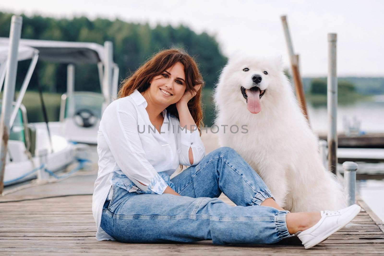 a happy woman with a big white dog sits on a pier by the sea at sunset by Lobachad