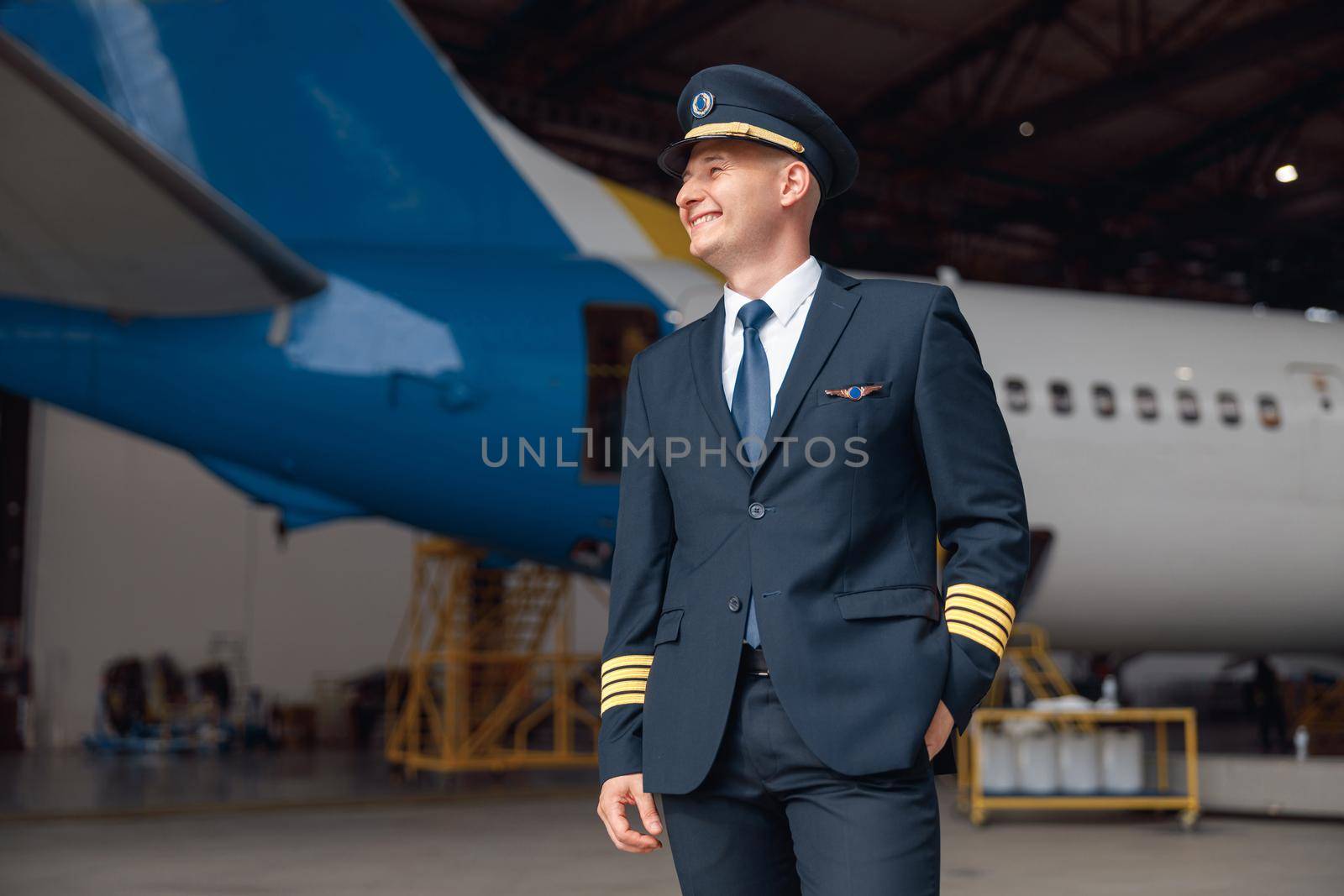 Cheerful pilot in uniform smiling away, standing in front of big passenger airplane in airport hangar. Aircraft, occupation, transportation concept