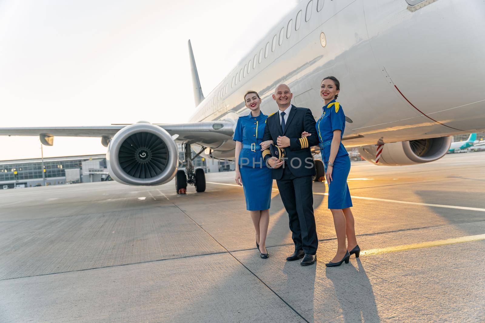Smiling male pilot in uniform posing together with two air stewardesses near airplane outdoor