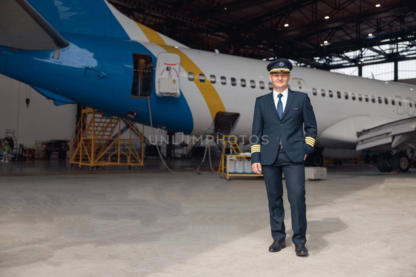 Full length shot of proud pilot in uniform smiling at camera, standing in front of big passenger airplane in airport hangar by Yaroslav_astakhov