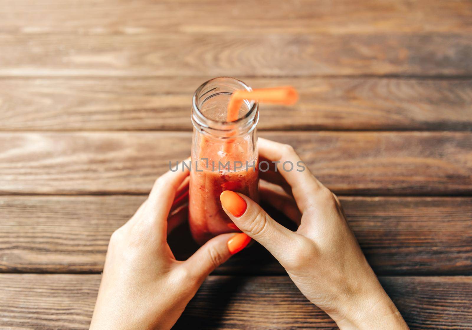 Woman’s hands with bright orange manicure holding bottle of fresh berry smoothie at wooden table. Point of view in first person.