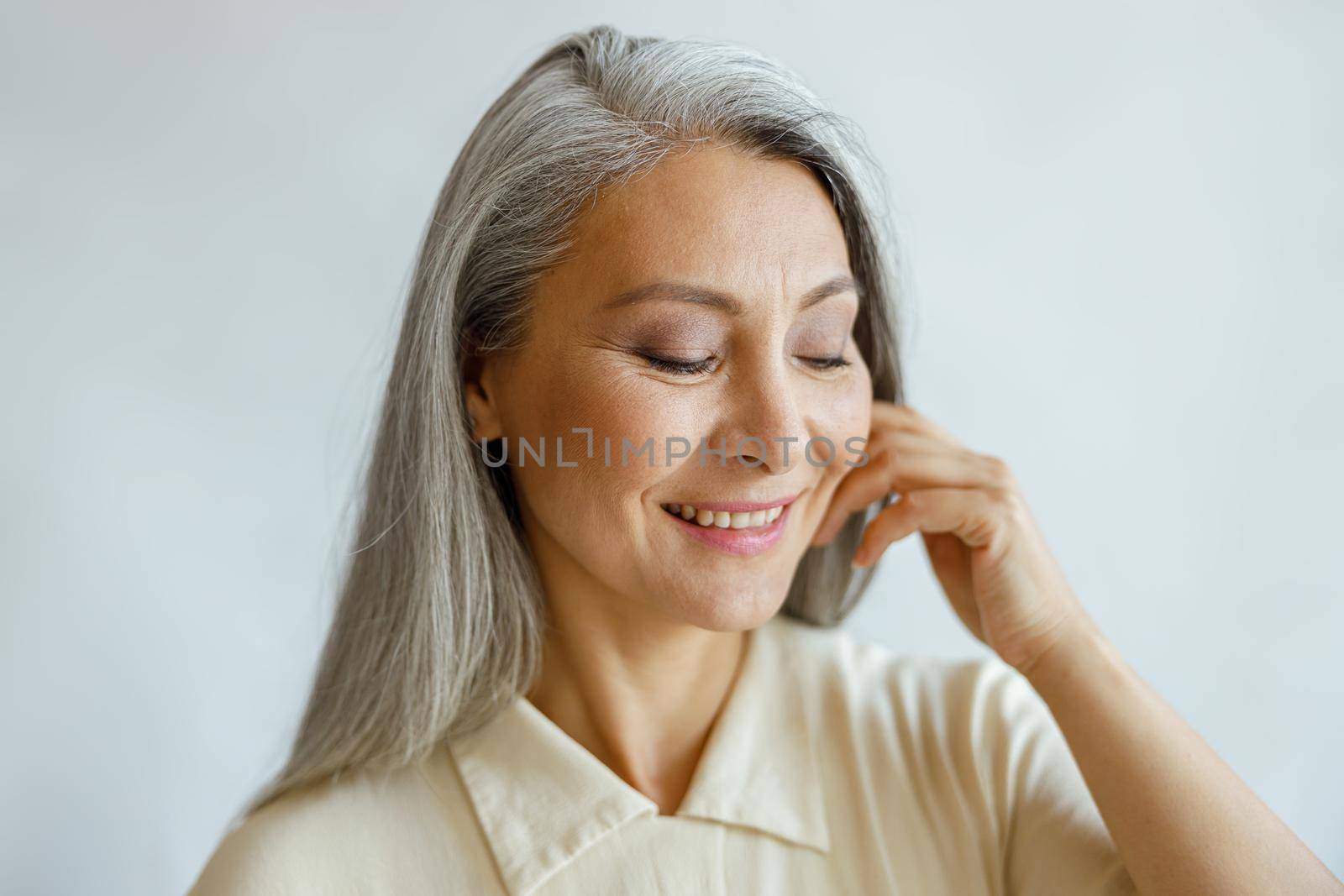 Attractive smiling grey haired lady in stylish shirt touches cheek by hand on light background in studio. Mature beauty lifestyle