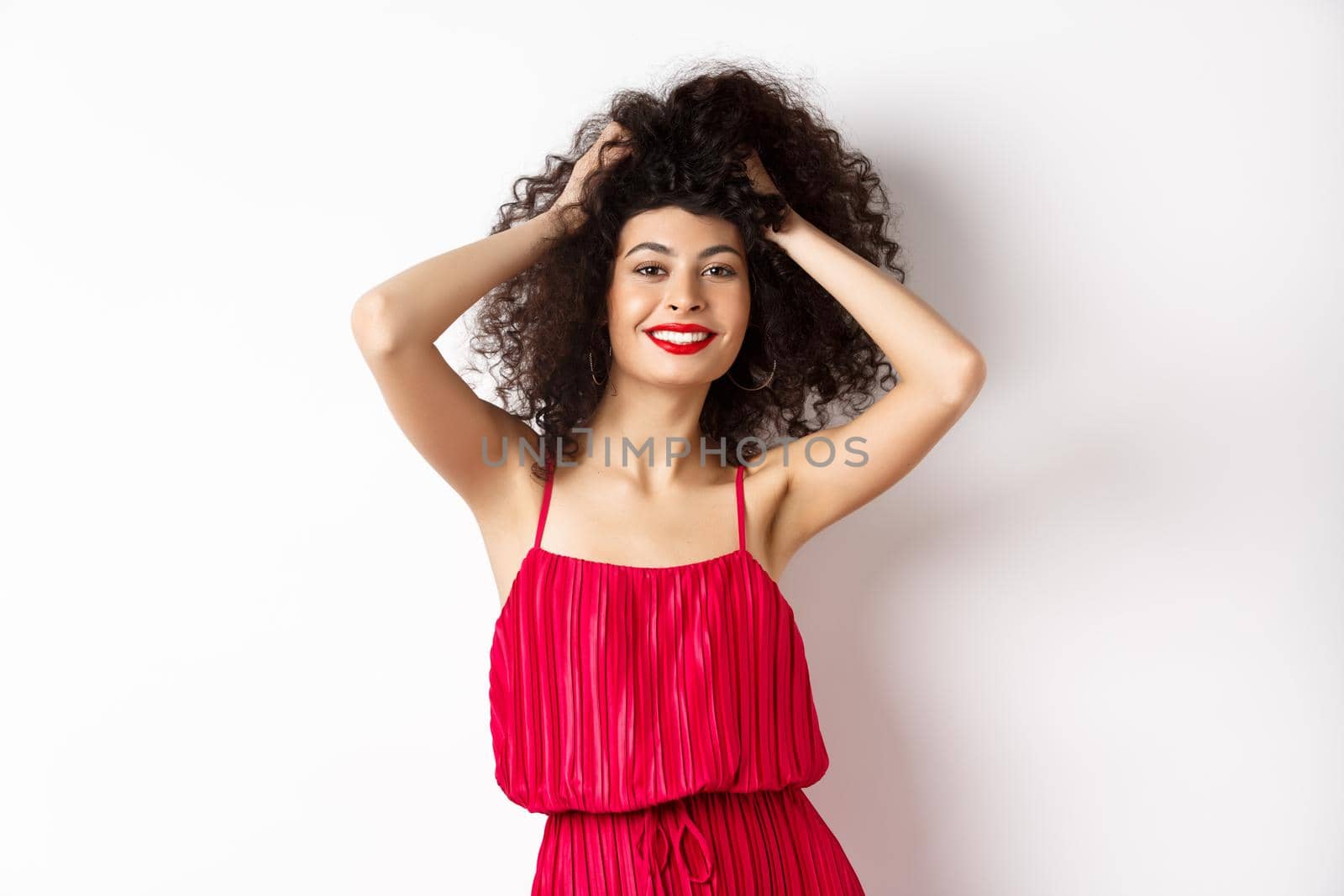 Beauty and fashion. Carefree woman in red dress and makeup, touching curly hair and smiling happy, standing on white background by Benzoix