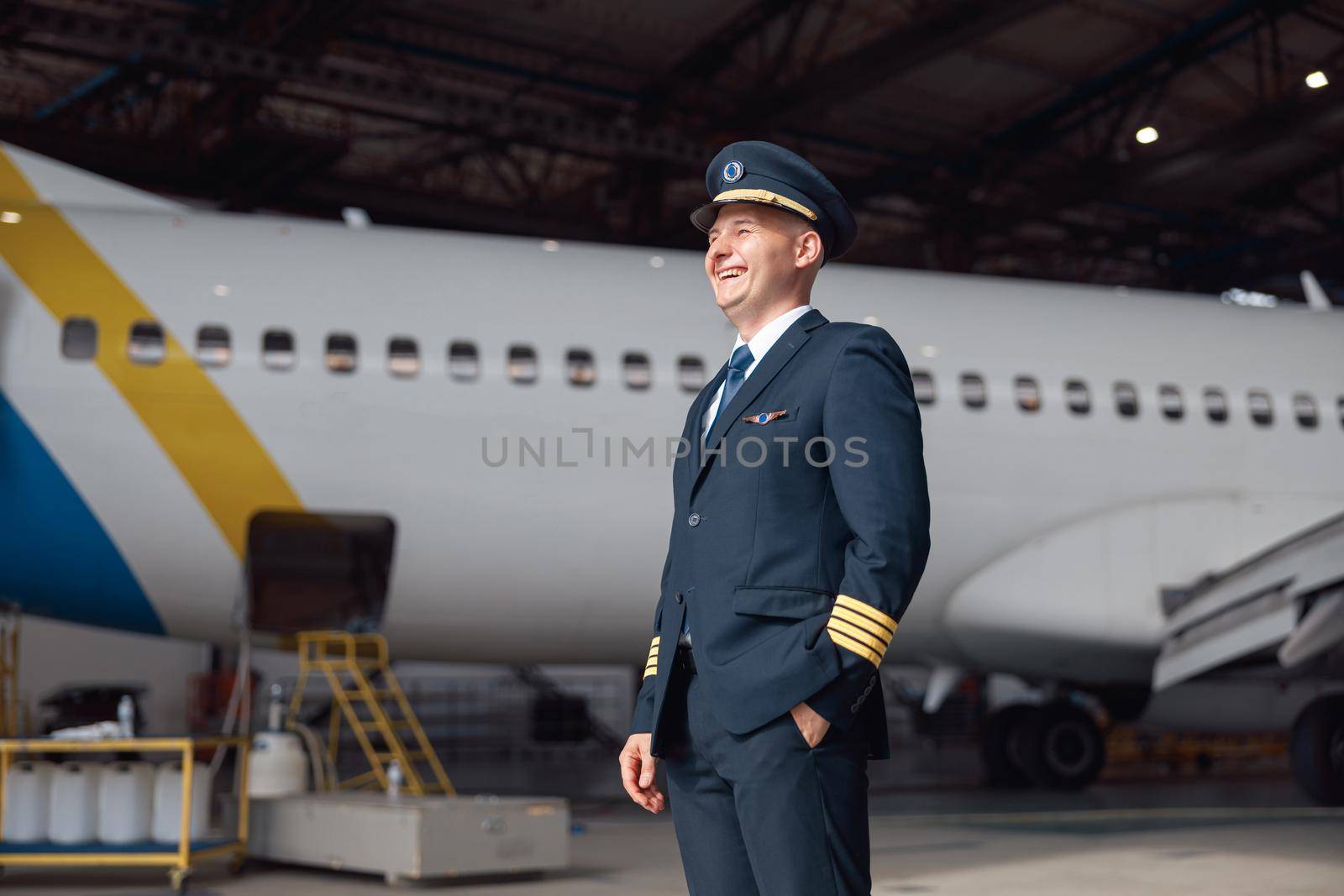 Portrait of happy pilot in uniform looking away, ready for flight, standing in front of big passenger airplane in airport hangar. Aircraft, occupation, transportation concept