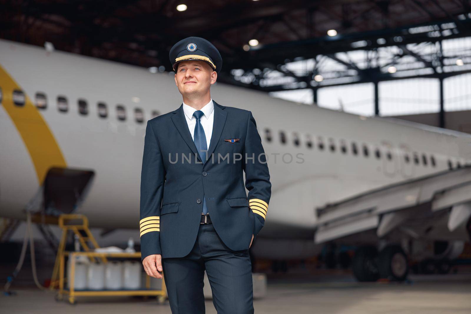 Thoughtful pilot in uniform looking away, standing in front of big passenger airplane in airport hangar. Aircraft, occupation, transportation concept