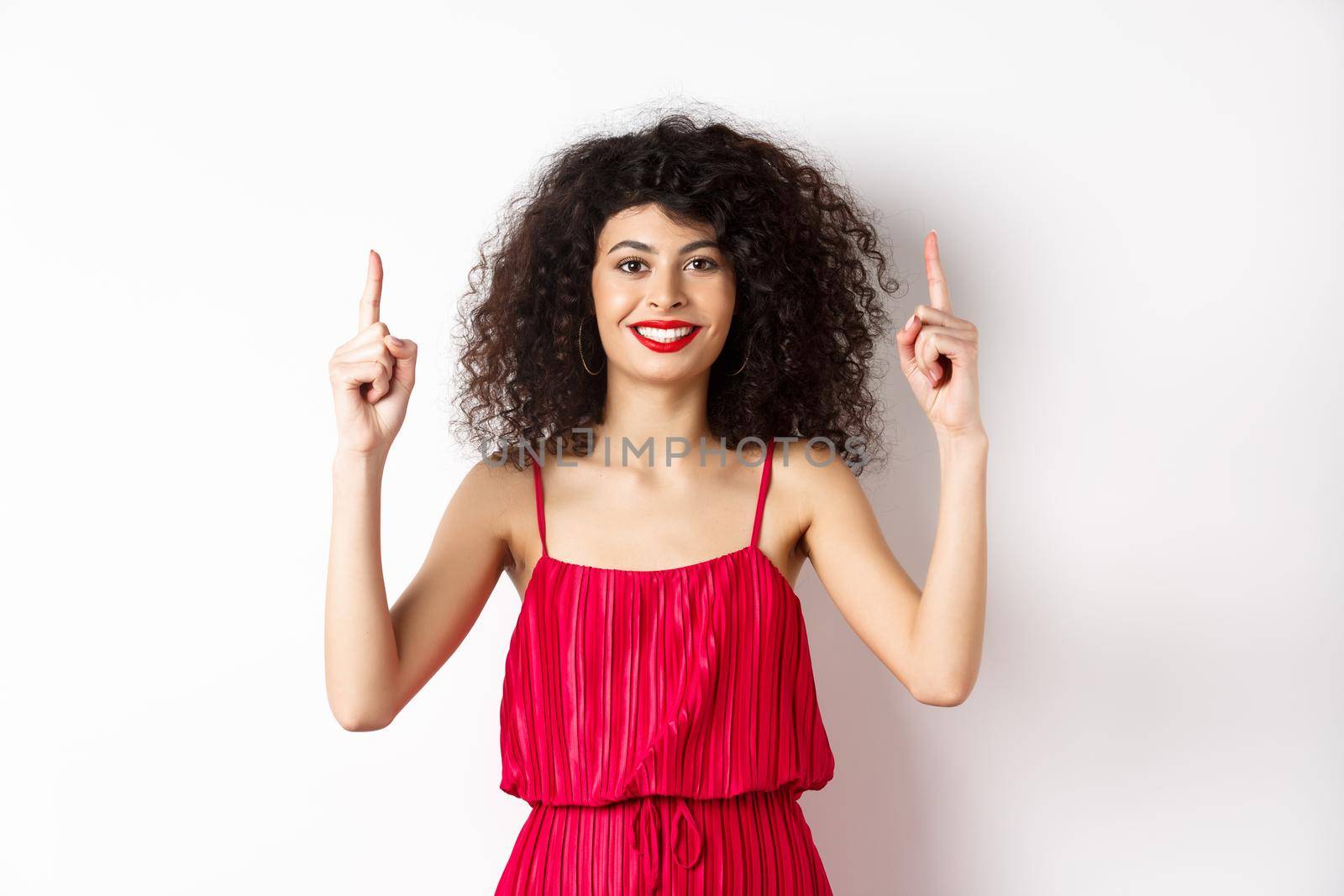 Smiling woman in elegant red dress and makeup, pointing fingers up and showing promo offer on valentines day, standing over white background by Benzoix